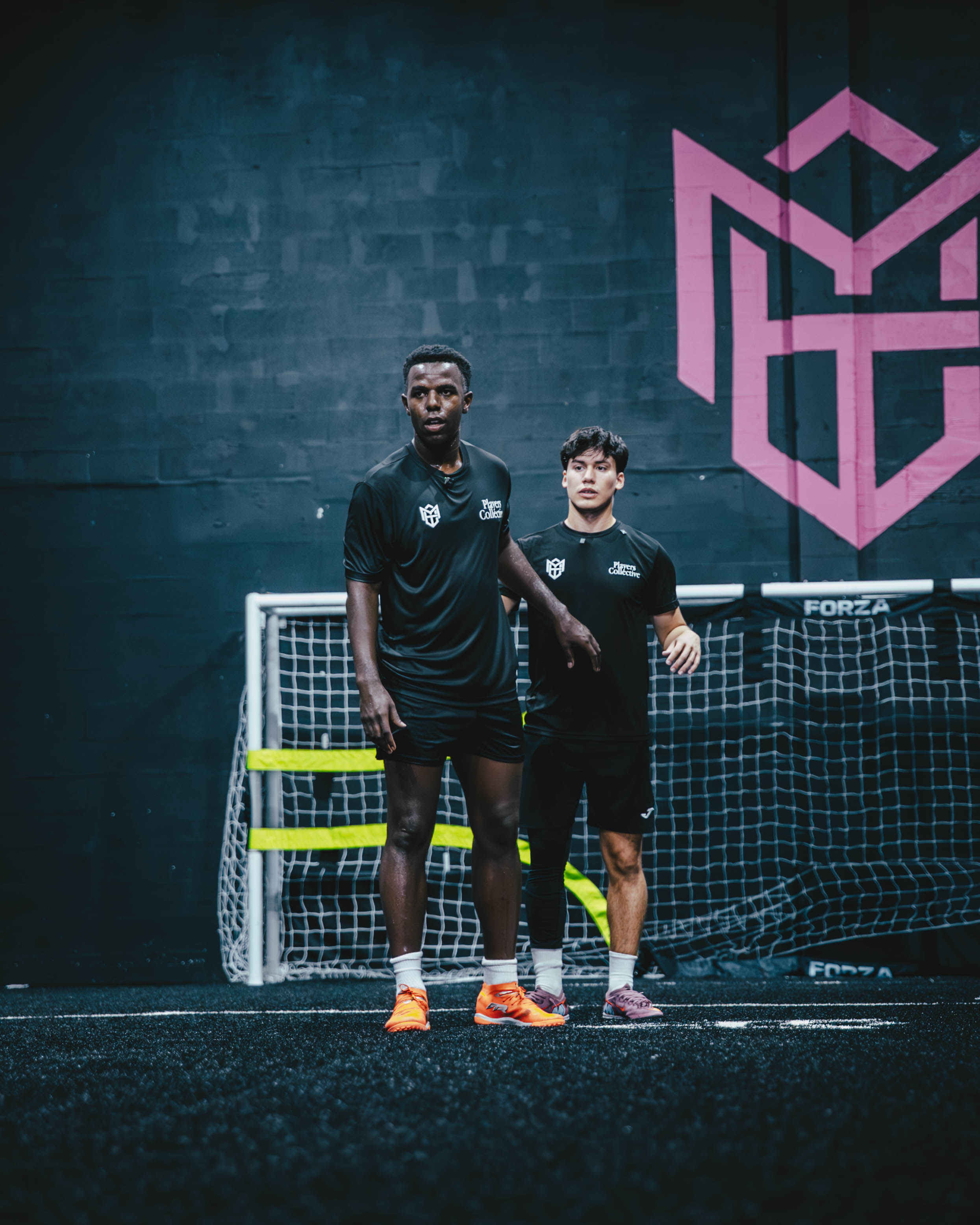 Two male soccer players in black uniforms standing in front of an indoor soccer goal with a pink geometric logo on the dark wall behind them.