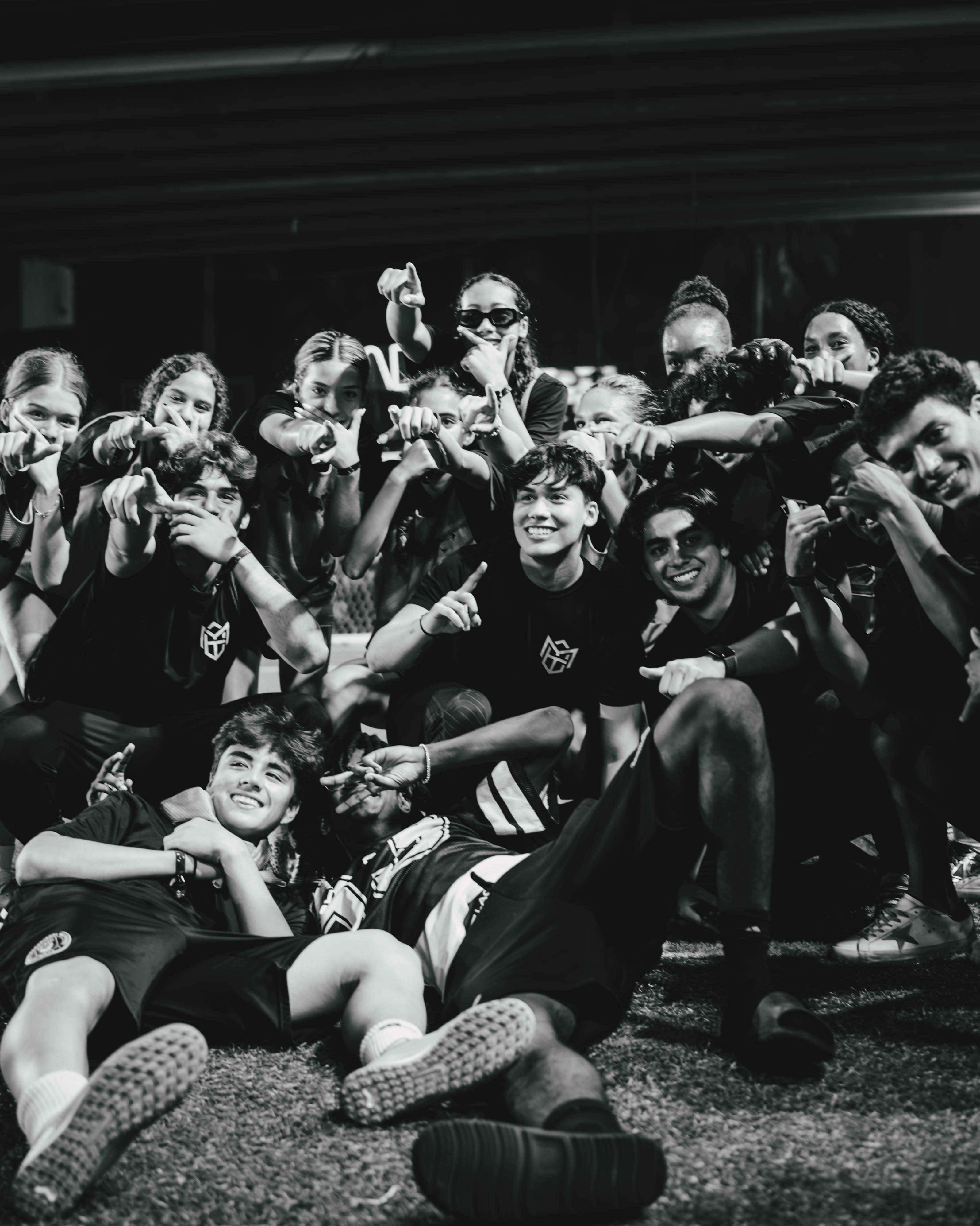 Group of young people in sportswear posing playfully on a sports field, some pointing towards the camera, smiling and lying on the ground.