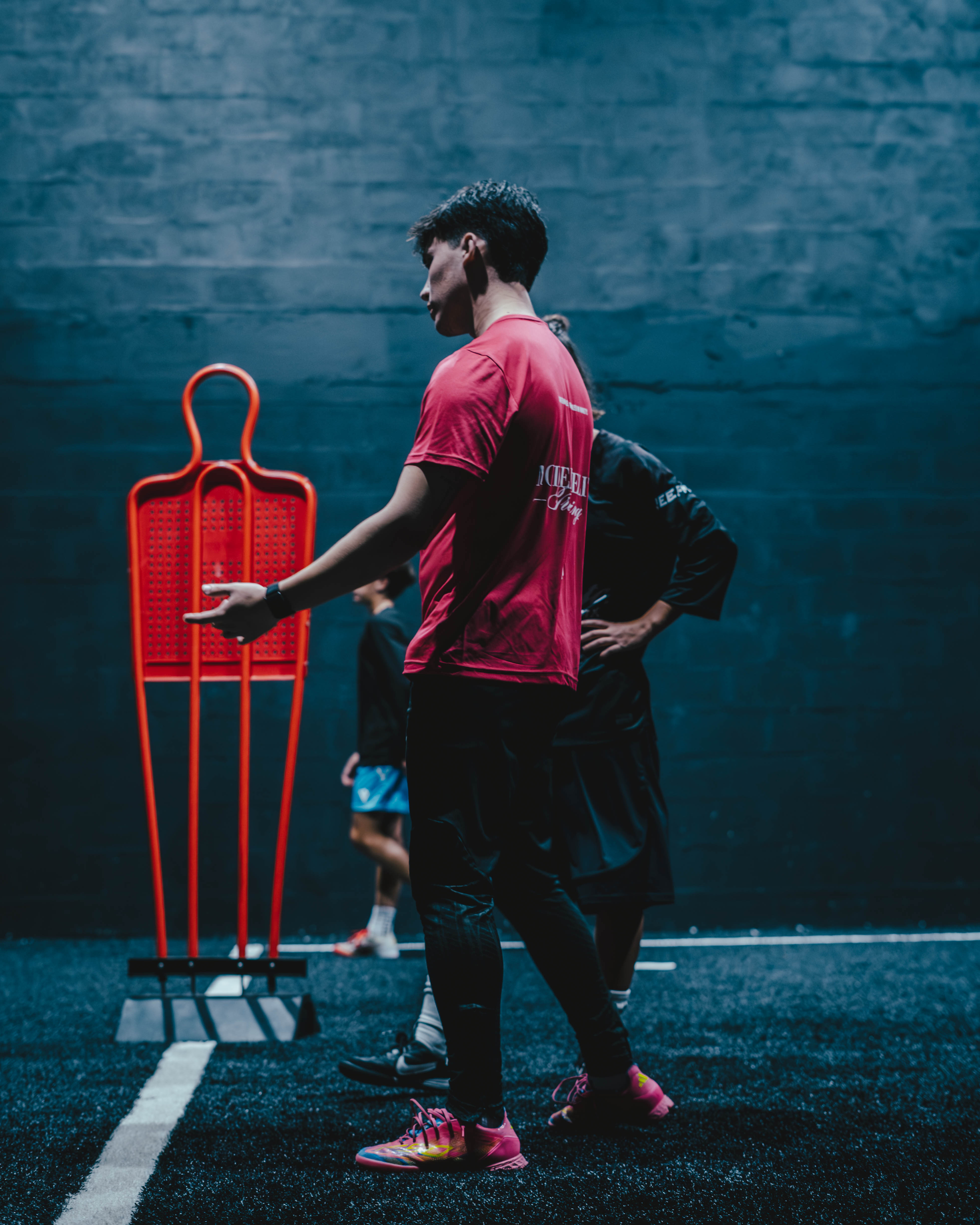 Two young men standing on a dark indoor sports field near a red training mannequin.