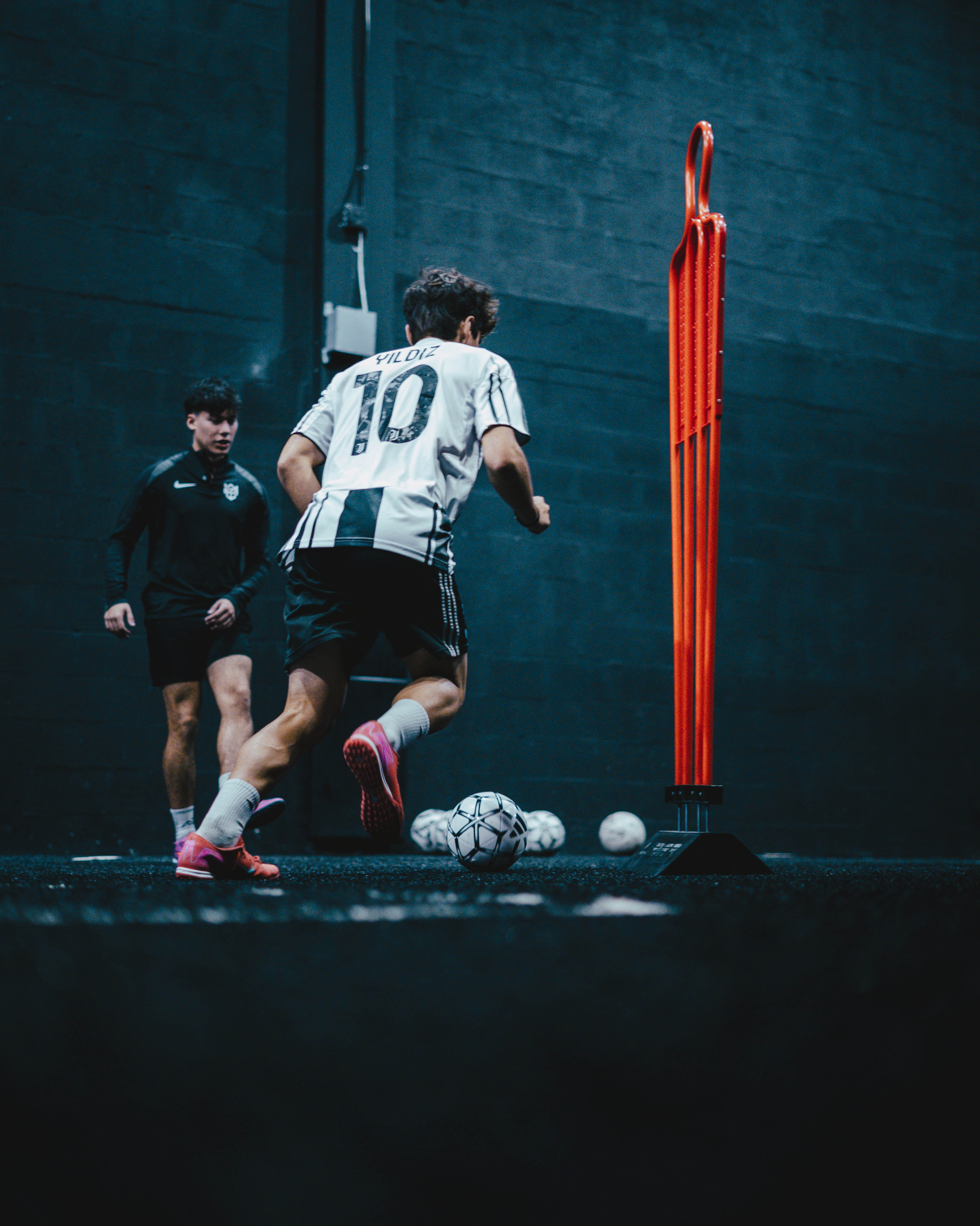 Two soccer players training indoors, one wearing a white and black jersey dribbling a soccer ball past an orange training mannequin.