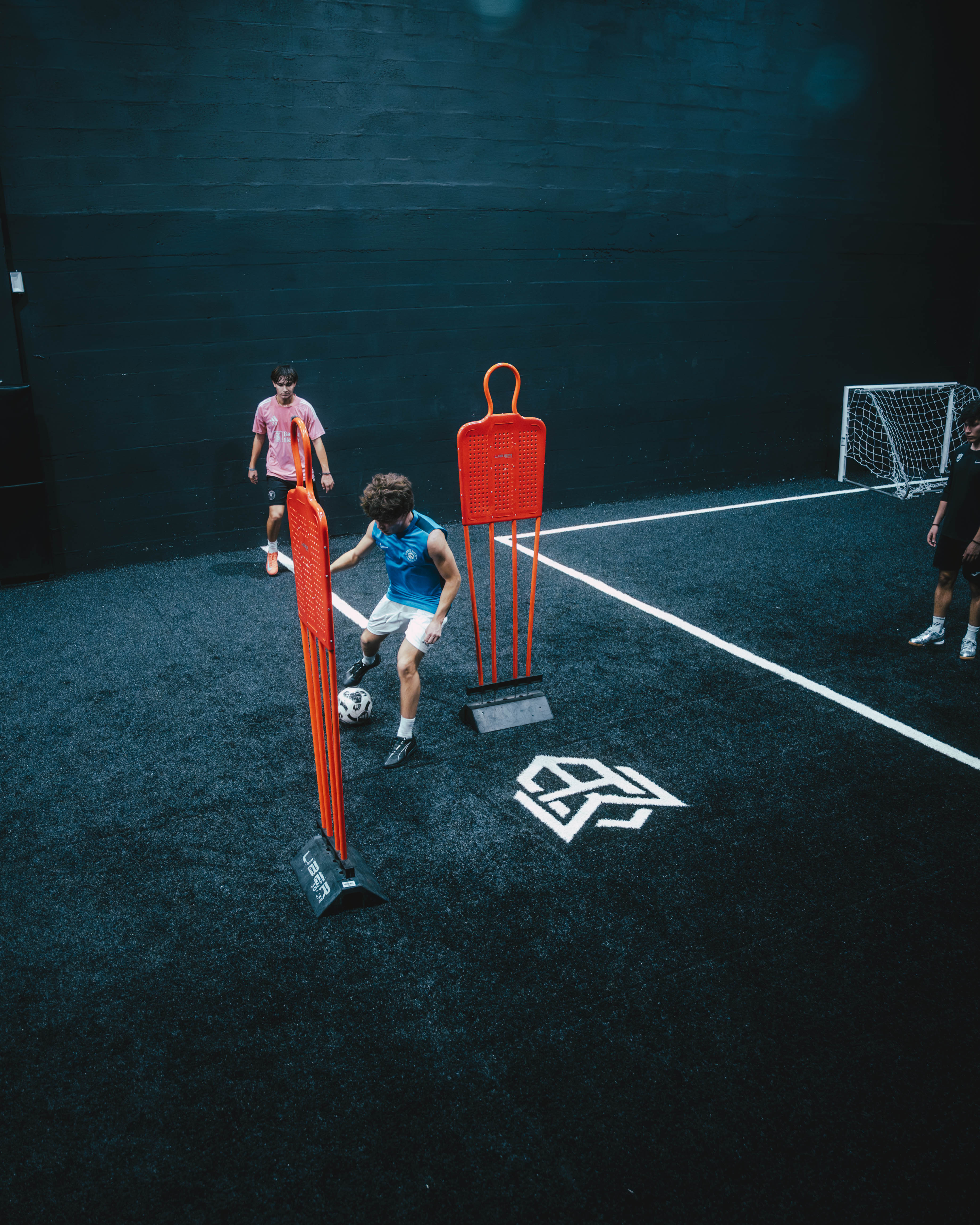 Three boys playing soccer on an artificial turf field with orange training mannequins and a small goal in the background.