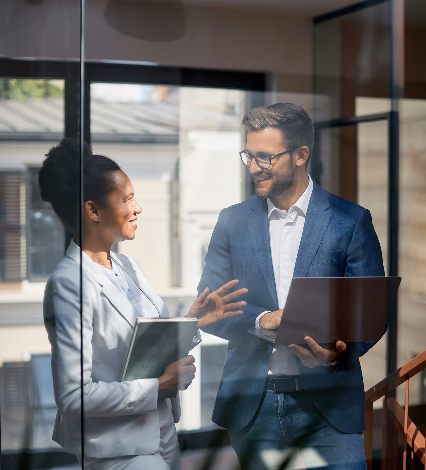 Two professionals discussing work on laptop in modern office setting near window