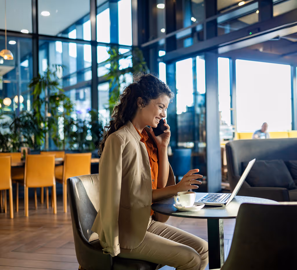 Woman on phone at laptop in modern office with plants and floor-to-ceiling windows