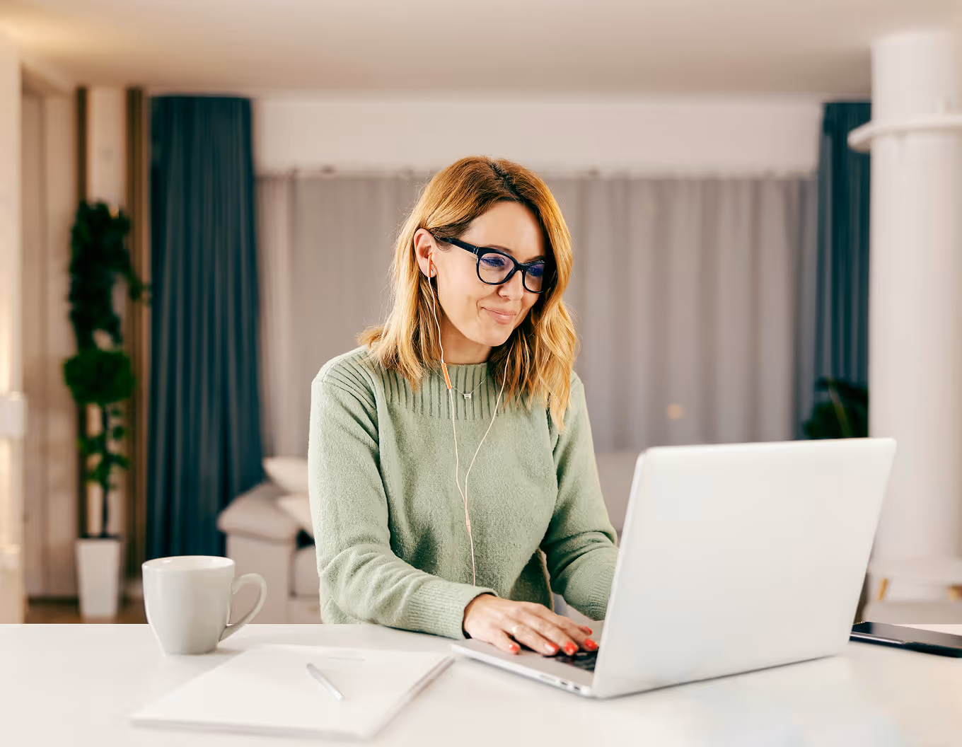 Woman with glasses wearing green sweater working on laptop at desk with coffee