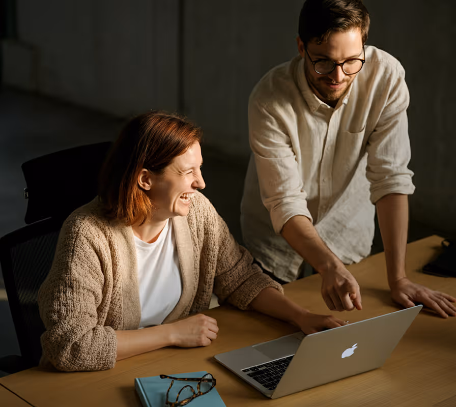 Two professionals collaborate at desk with laptop, smiling while working together.