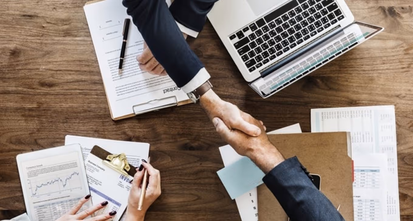 Two business professionals shaking hands over wooden desk with laptop and financial documents.