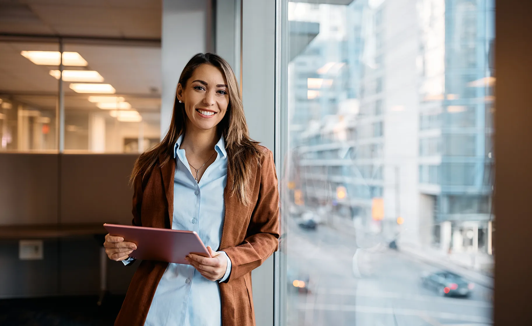 Professional woman smiling at camera holding tablet in modern office with city view
