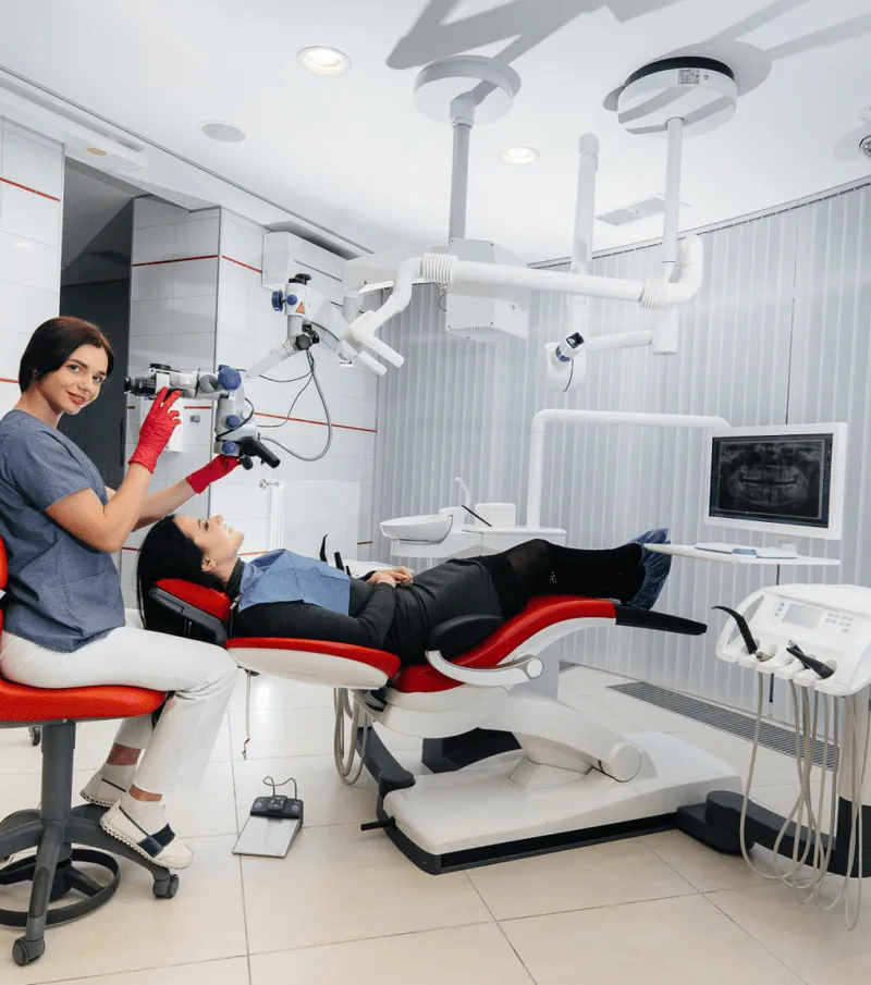 Dentist in red gloves adjusting a microscope while a patient reclines on a red and white dental chair in a modern dental clinic.