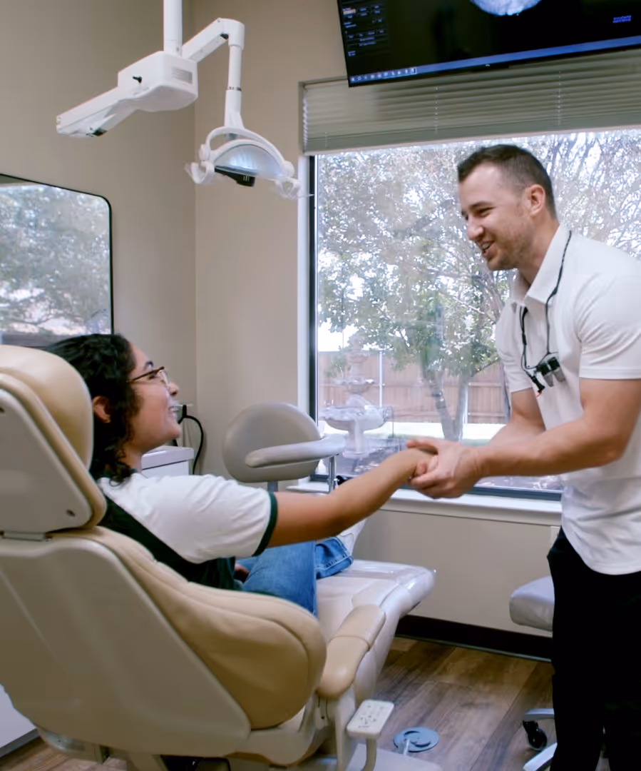 Dentist shaking hands with a smiling patient seated in a dental chair in a clinic room with large window.