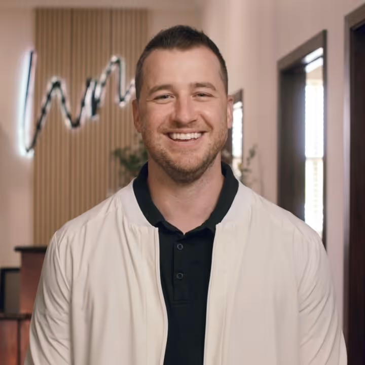 Smiling man with short hair wearing a white jacket over a black collared shirt, standing indoors with a modern decor background.