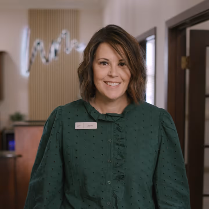 Smiling woman with shoulder-length brown hair wearing a green blouse with a name tag in an indoor office setting.