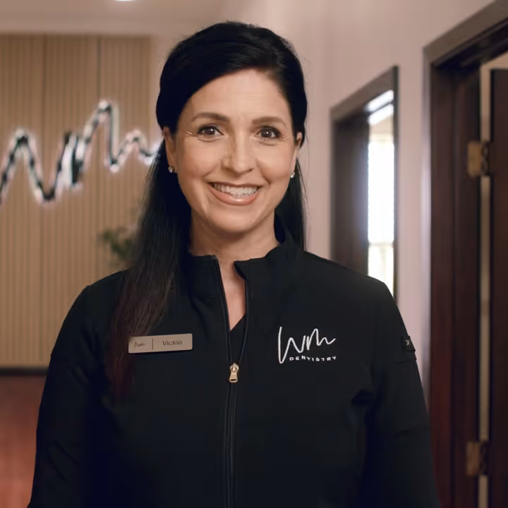 Smiling woman with dark hair wearing a black jacket with 'WM Dentistry' logo and a name tag reading 'Vickie'.