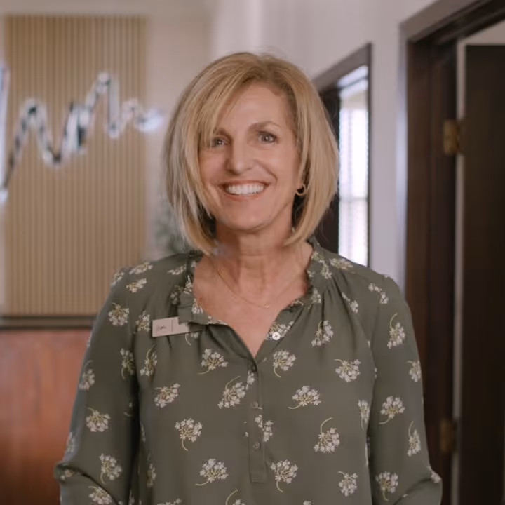Smiling woman with short blonde hair wearing a green floral blouse standing indoors.