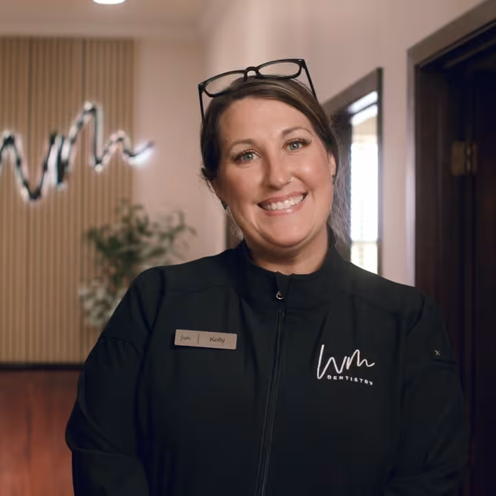 Smiling woman wearing black WM Dentistry uniform with glasses on her head in an indoor setting.