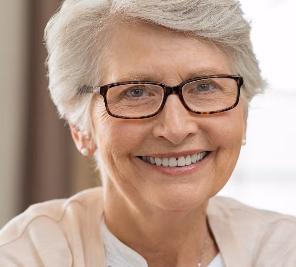 Smiling elderly woman with short gray hair and glasses wearing a light-colored top.