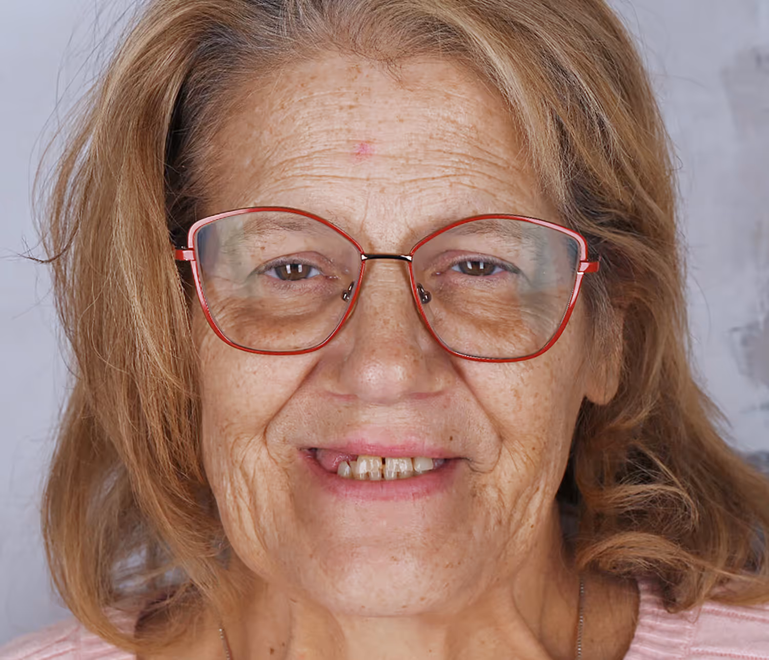 Close-up of an older woman with light brown hair, wearing red-framed glasses and smiling slightly with visible worn teeth.