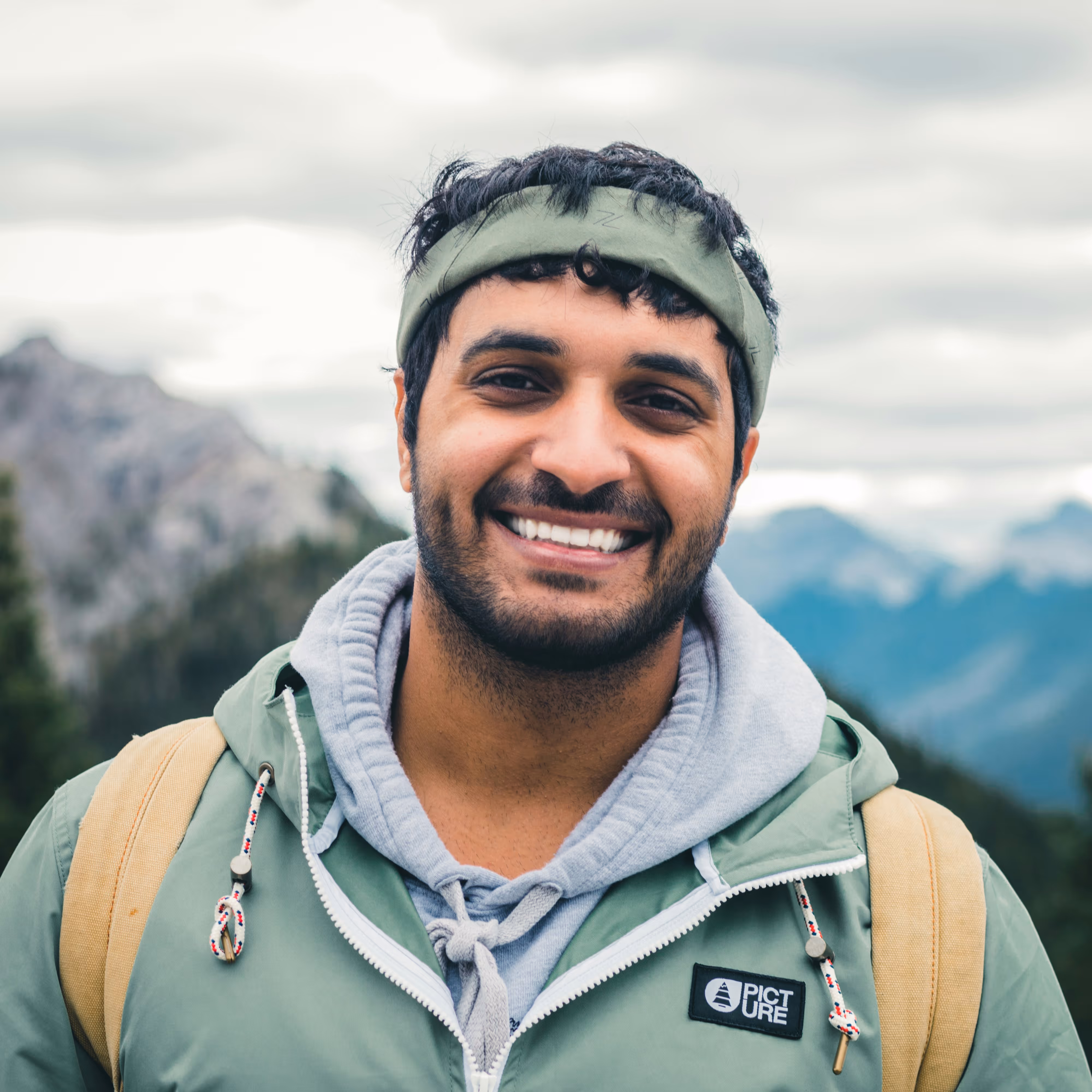 Smiling man with headband and backpack outdoors with mountains and cloudy sky in the background.
