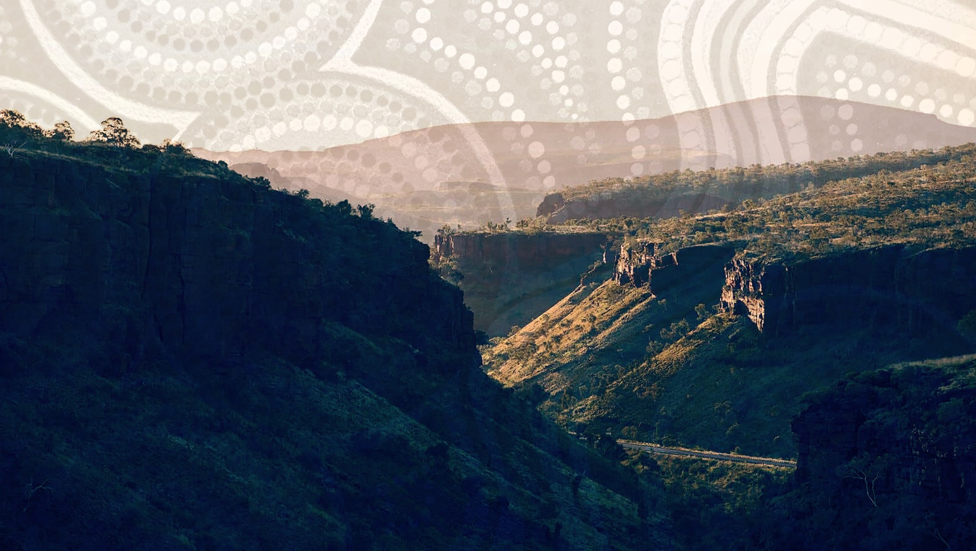 Mountain valley with shadows and sunlight, trees on ridges, and subtle Aboriginal dot art overlay.