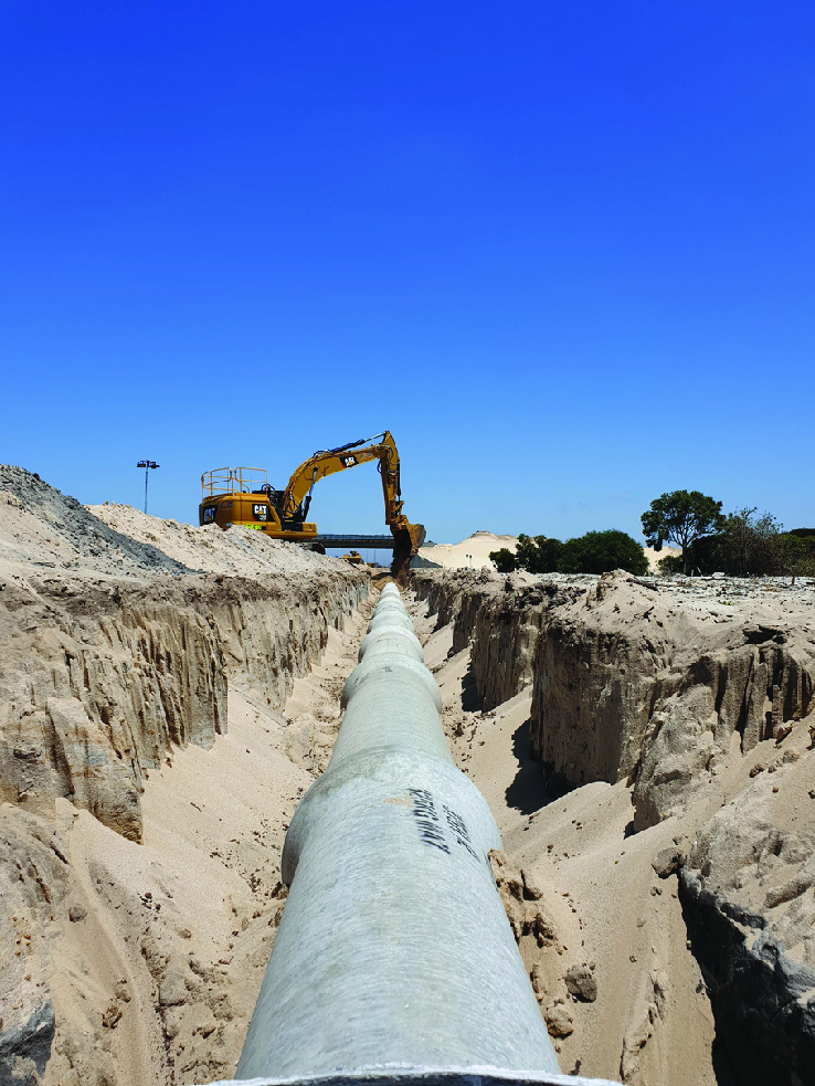 Long concrete pipeline laid in a trench with a yellow excavator in the background under clear blue sky.
