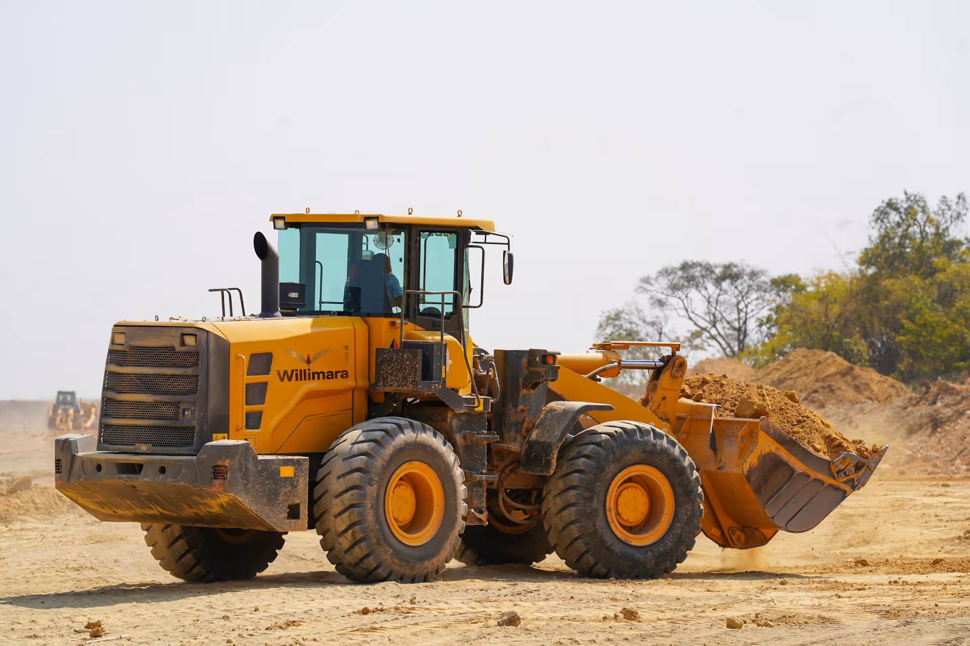 Yellow Willimara front loader heavy machinery moving dirt at a construction site with trees in background.