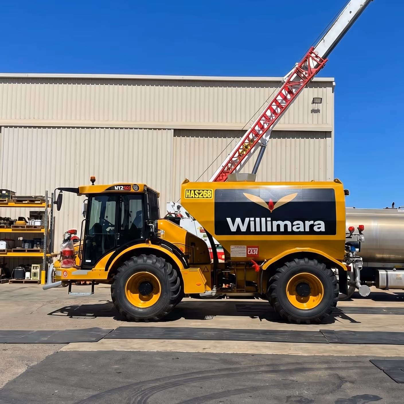 Yellow Willimara agricultural vehicle with large tires parked near a warehouse under a clear blue sky.