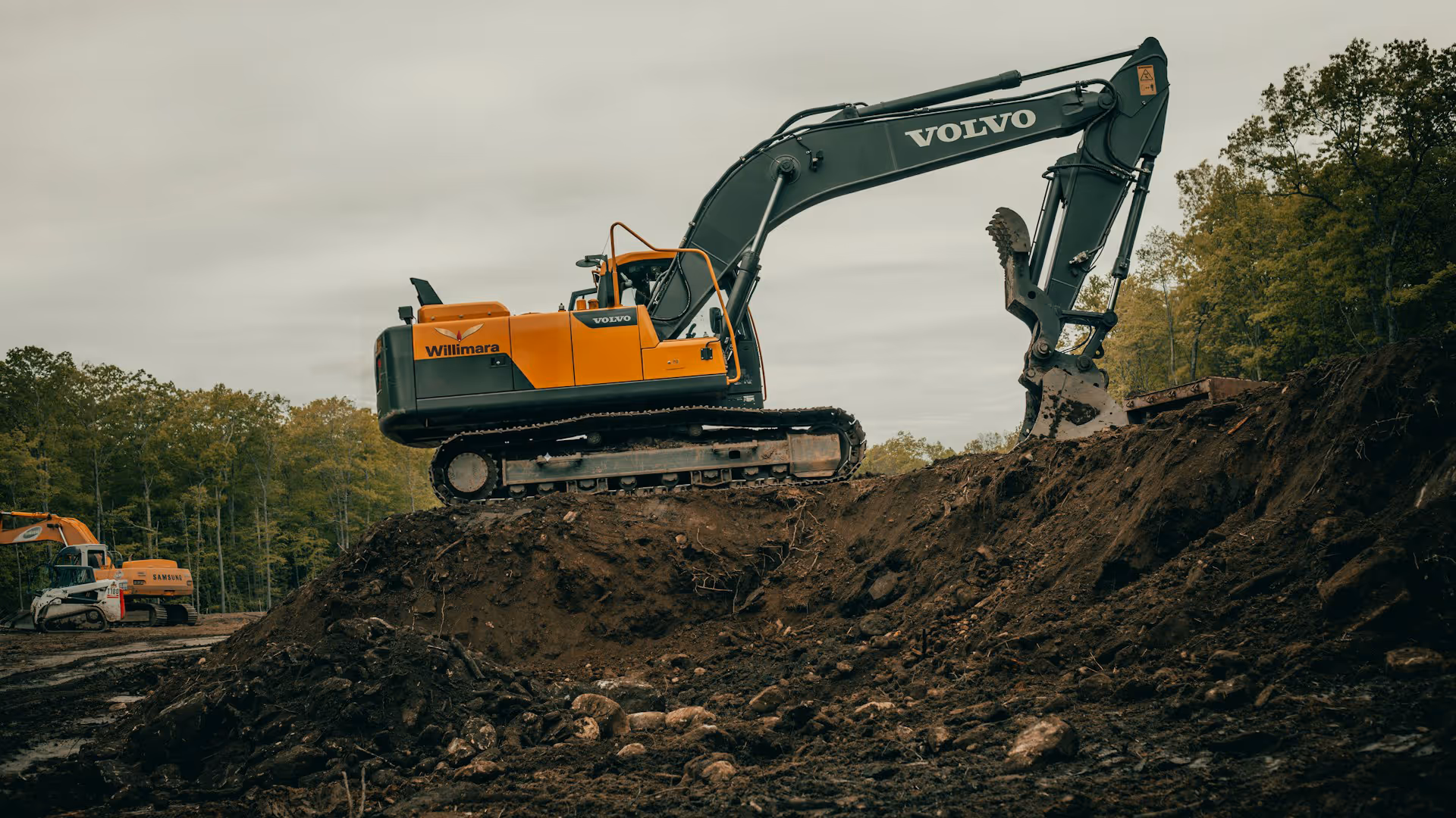 Yellow and black Volvo excavator with Willimara branding digging dirt in a forest clearing.