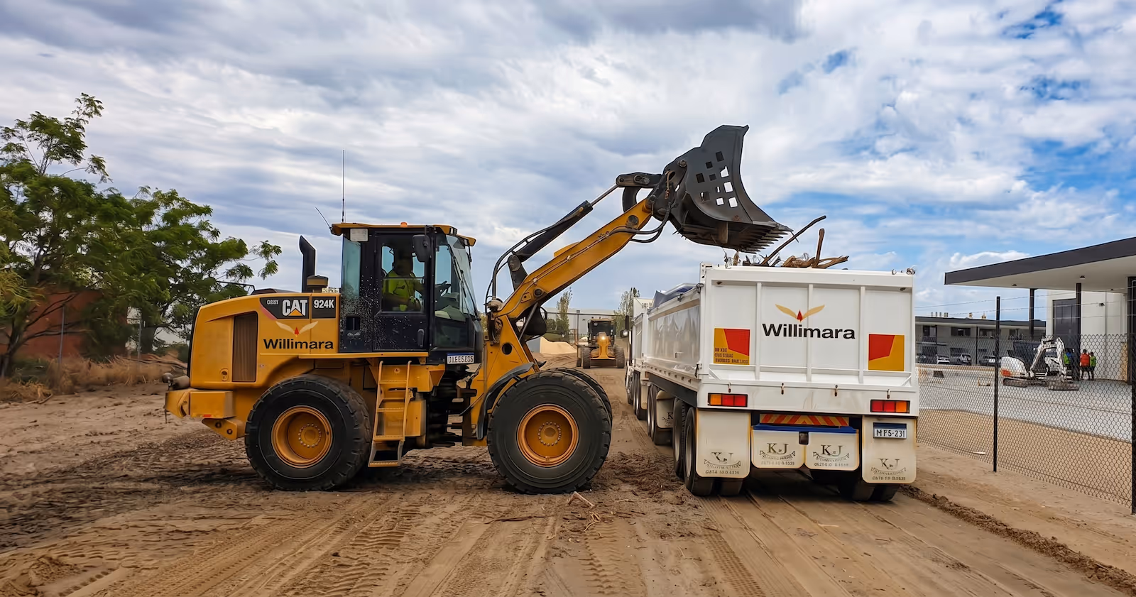 Yellow loader with Willimara logo loading debris into a white Willimara truck on a dirt road.