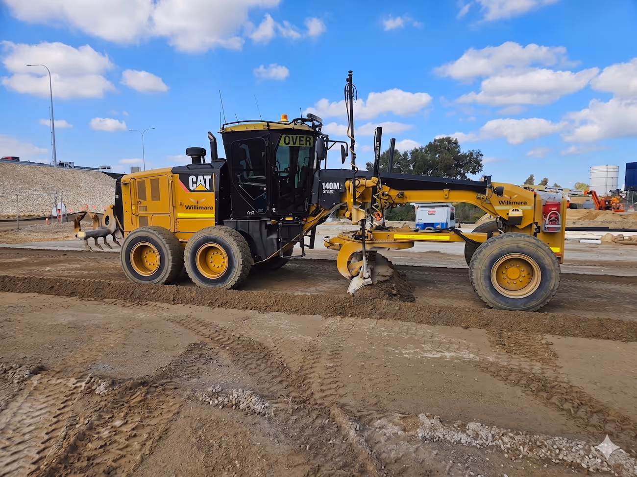 Yellow CAT 140M motor grader leveling dirt at a construction site under a blue sky.