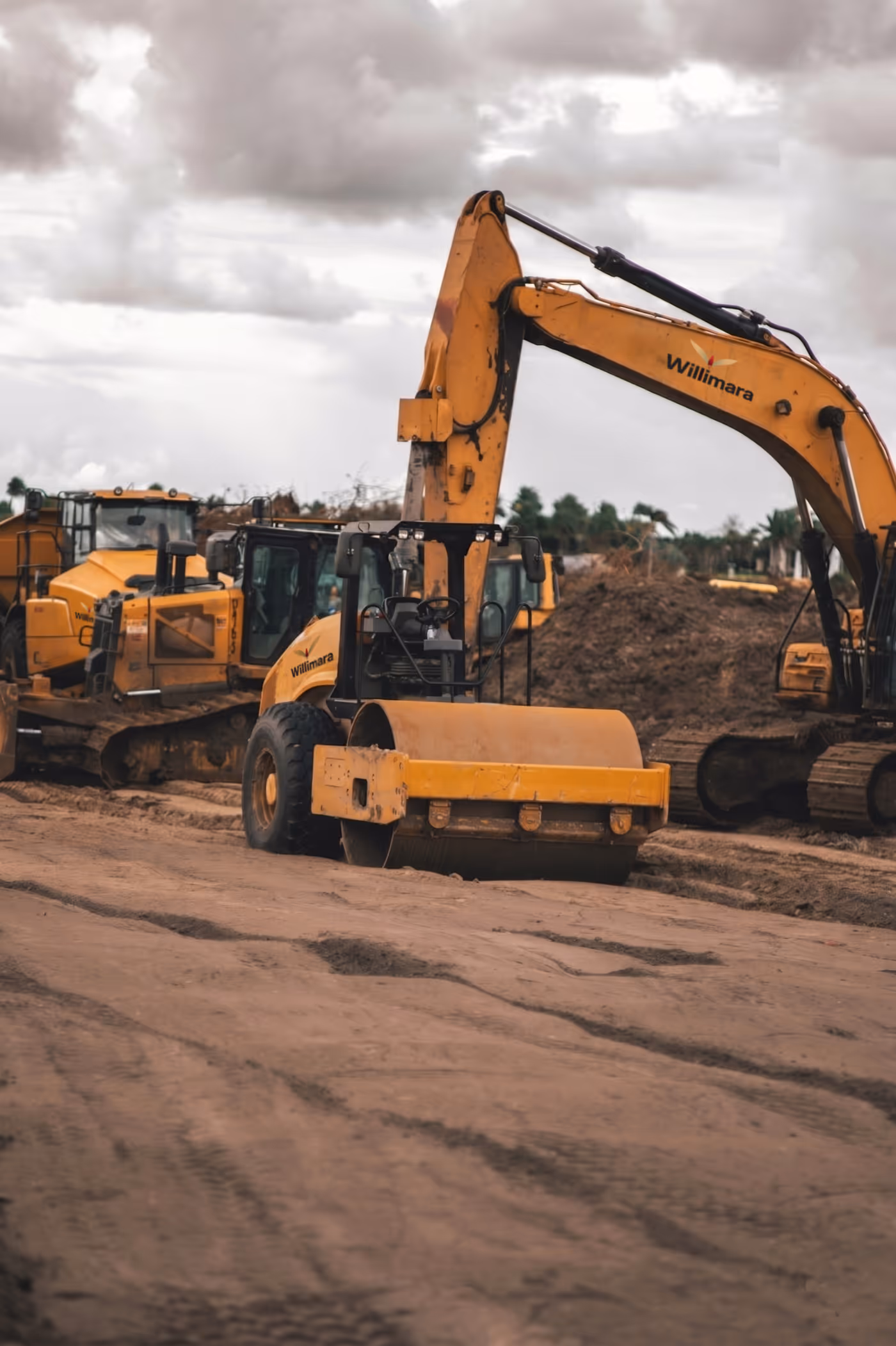 Yellow heavy construction machinery at a dirt site under cloudy sky, including excavators and a roller.