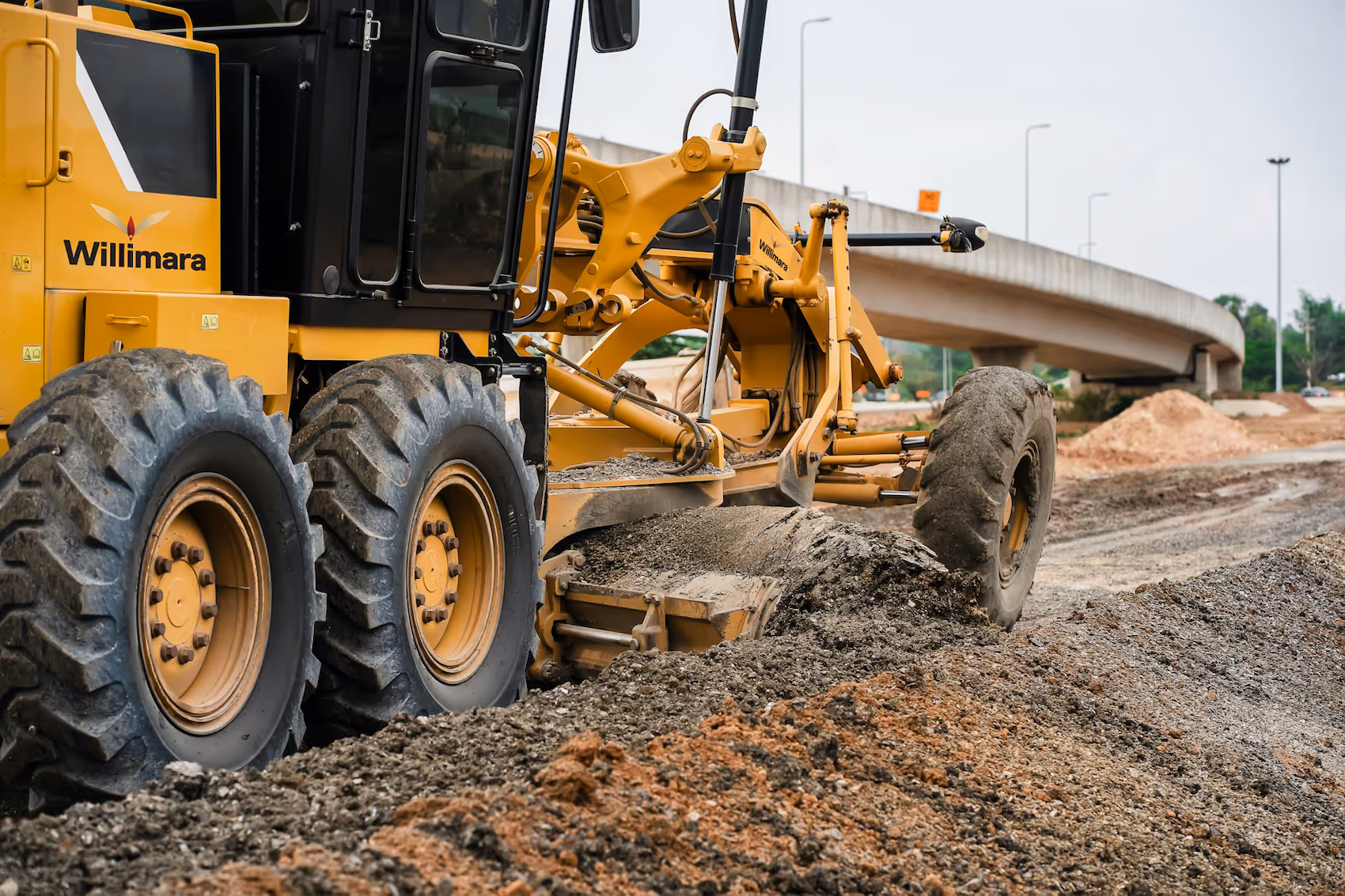 Yellow Willimara grader leveling soil on a construction site near a concrete bridge.