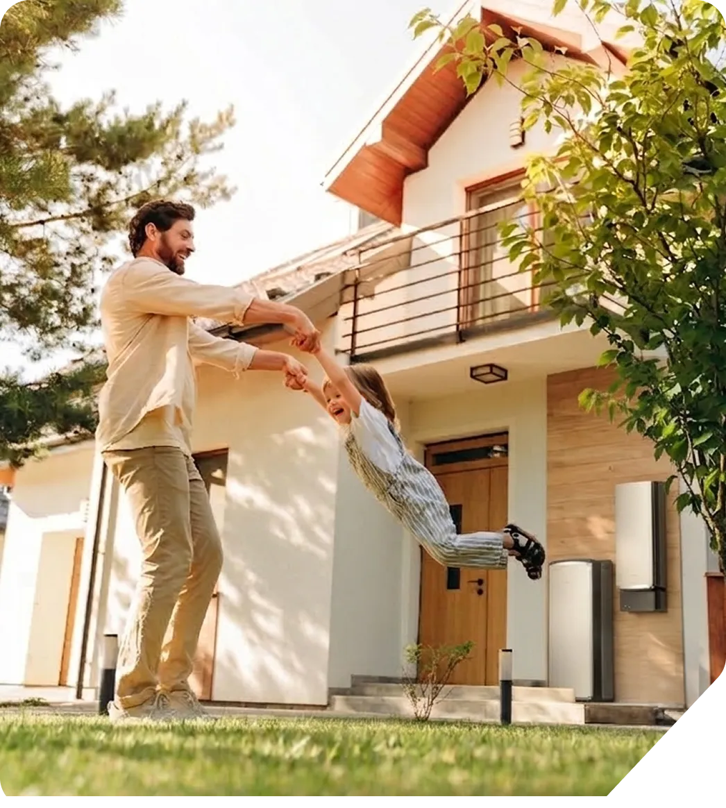 Man swinging a smiling child by the hands in front of a modern house with green lawn and trees.