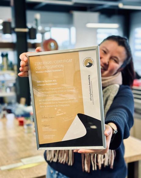 Foto von 15 Personen, die jeweils ein Diplom vor einem weißen Zelt im Freien halten.