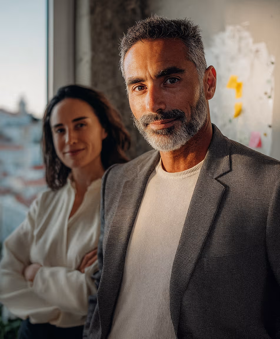 Confident man with gray beard in a blazer and woman in white blouse standing behind him near a window.