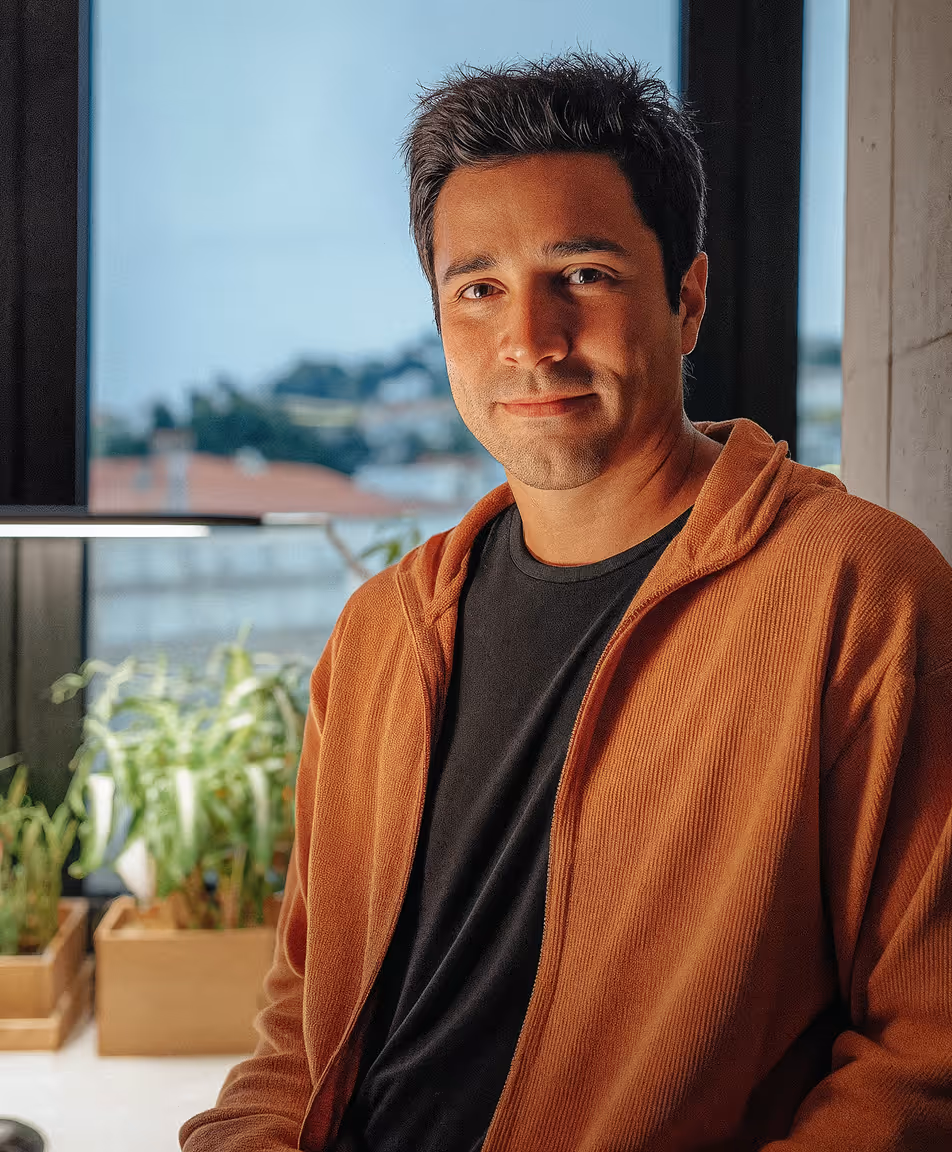 Man wearing a burnt orange hoodie and black shirt standing indoors with potted plants and a window in the background.