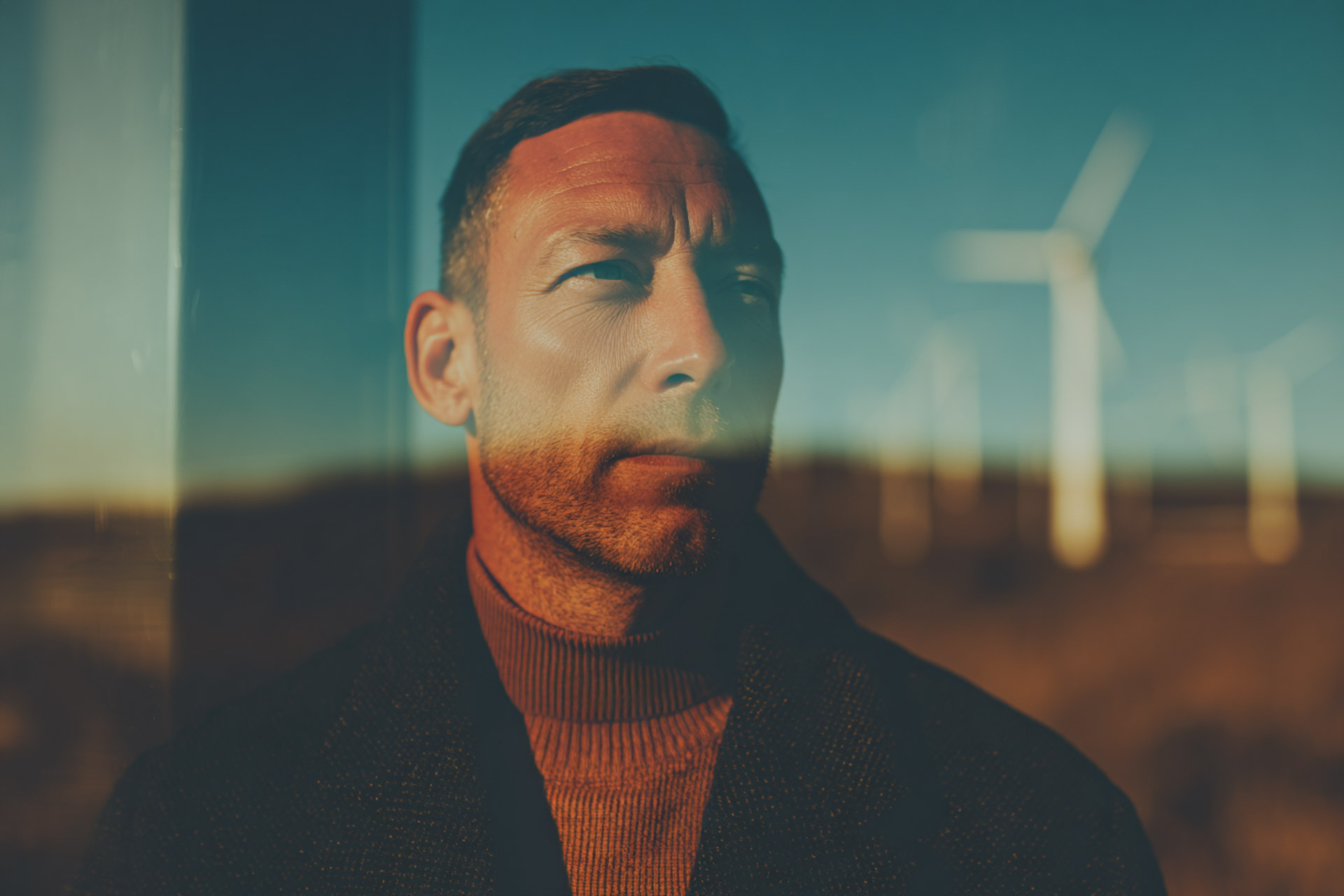 Close-up of a man with short hair and a beard looking sideways, with wind turbines blurred in the background.