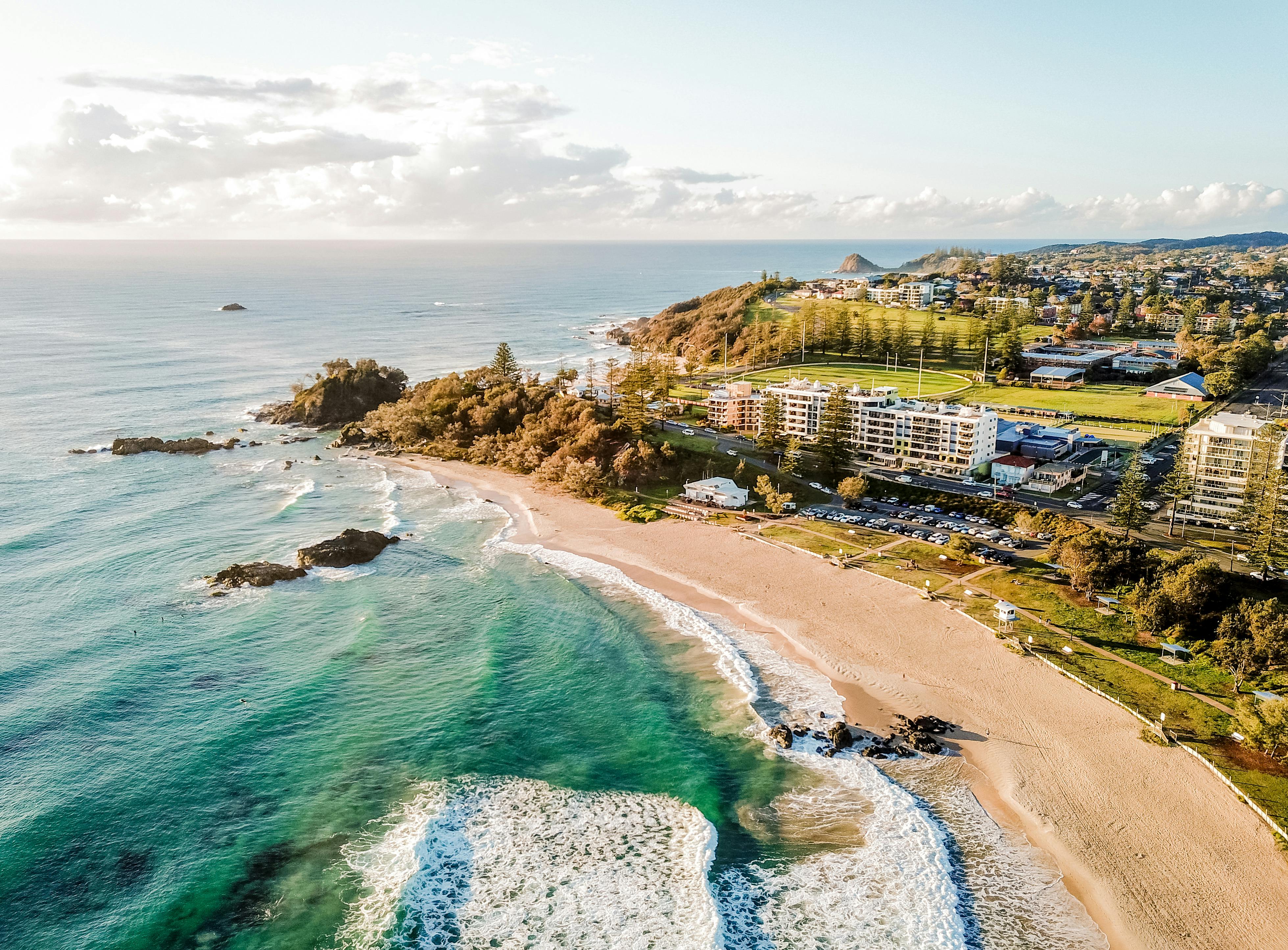 Aerial view of a Town Beach in Port Macquarie with clear turquoise water, waves, rocky outcrops, and a nearby town with buildings and green fields.