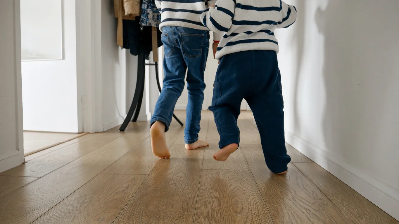 Two young children wearing striped sweaters and pants walking barefoot on a wooden floor hallway.