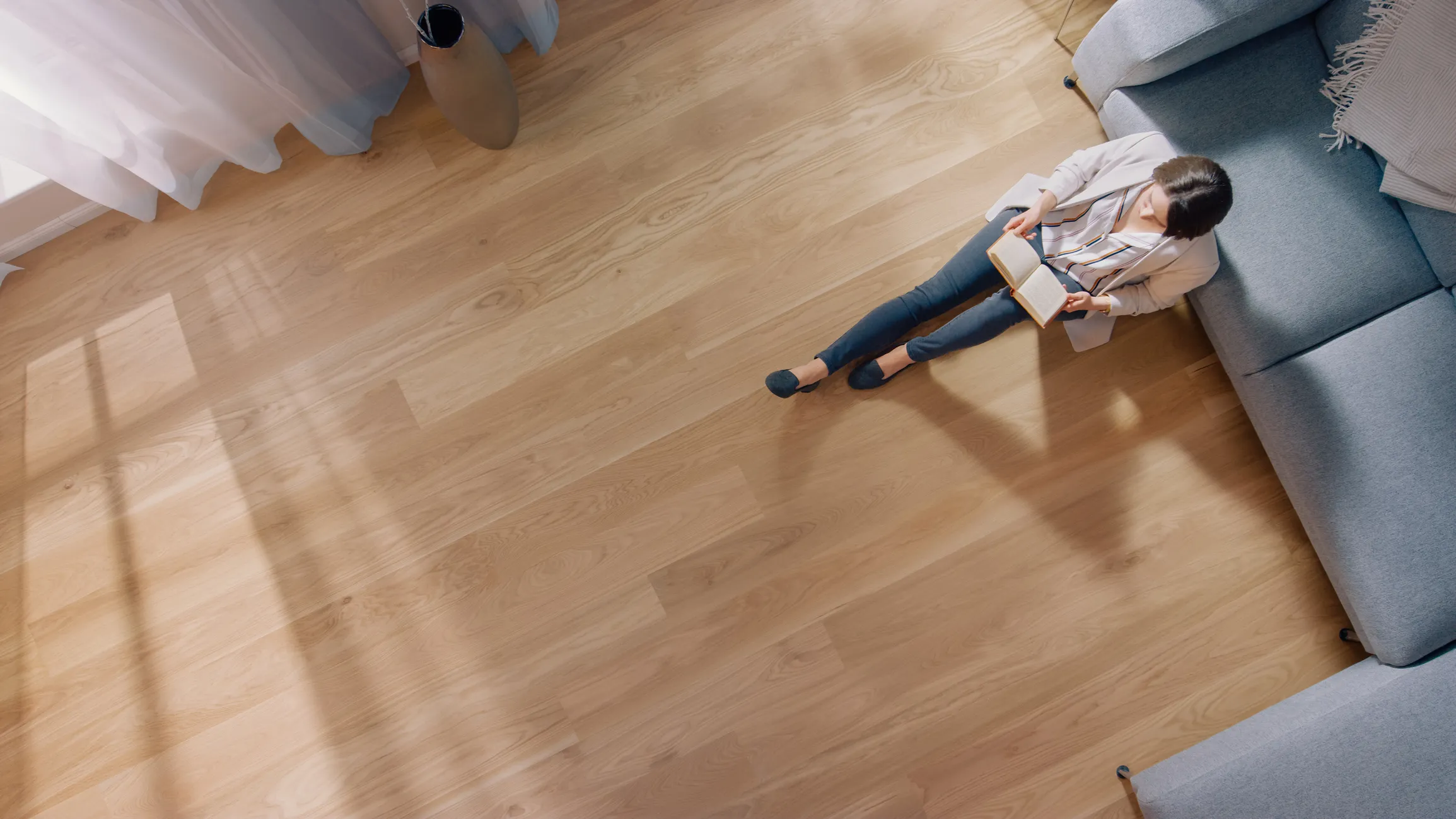 Woman sitting on a wooden floor next to a gray sofa, reading a book in natural light.