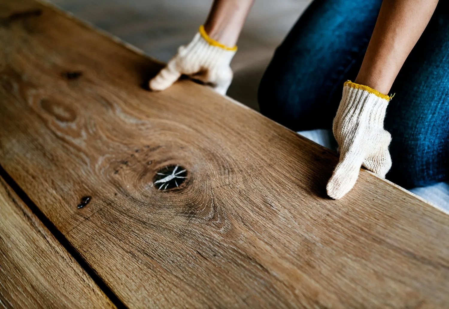 Hands wearing white work gloves holding a plank of wood with visible grain and a knot.