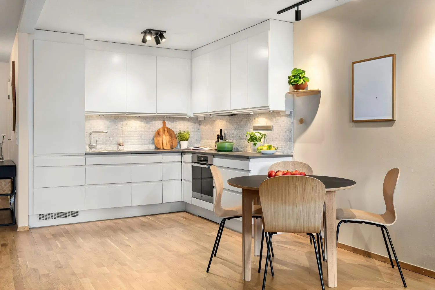 Modern kitchen with white cabinets, wooden floor, round dining table with four chairs, and green plants on the countertop.