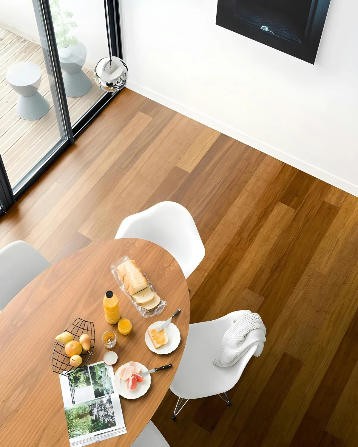 Top view of a wooden dining table with breakfast items including sliced bread, orange juice, fruit, and an open magazine, surrounded by white chairs on wooden floor beside glass doors.