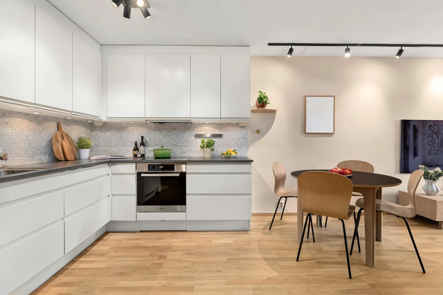 Modern kitchen with white cabinets, black countertop, built-in oven, dining table with four wooden chairs, and wooden floor.