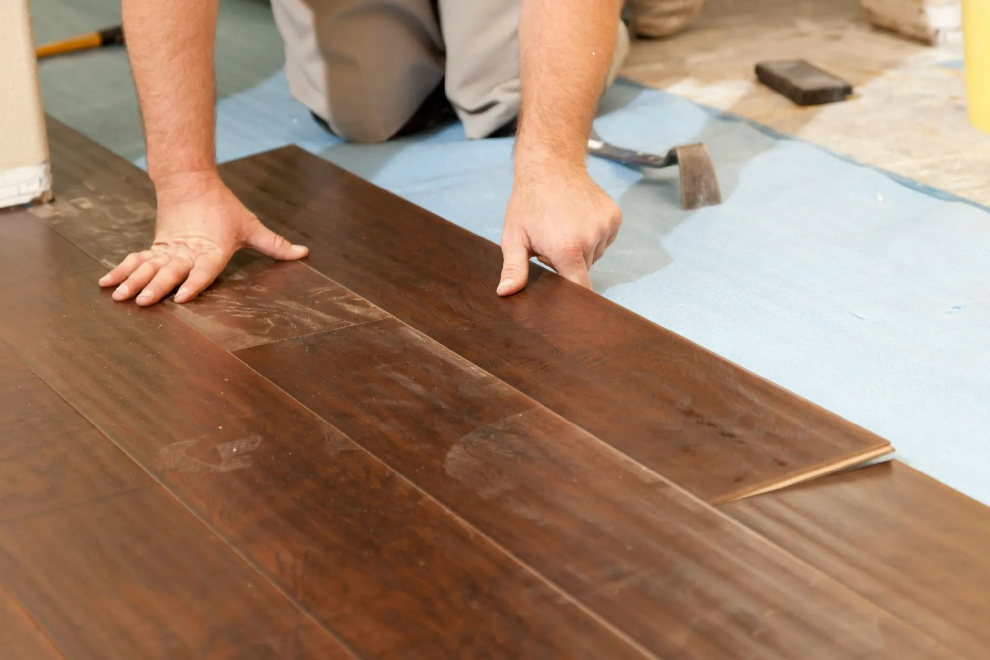 Person installing dark wood laminate flooring with hands placing a plank on blue underlayment.