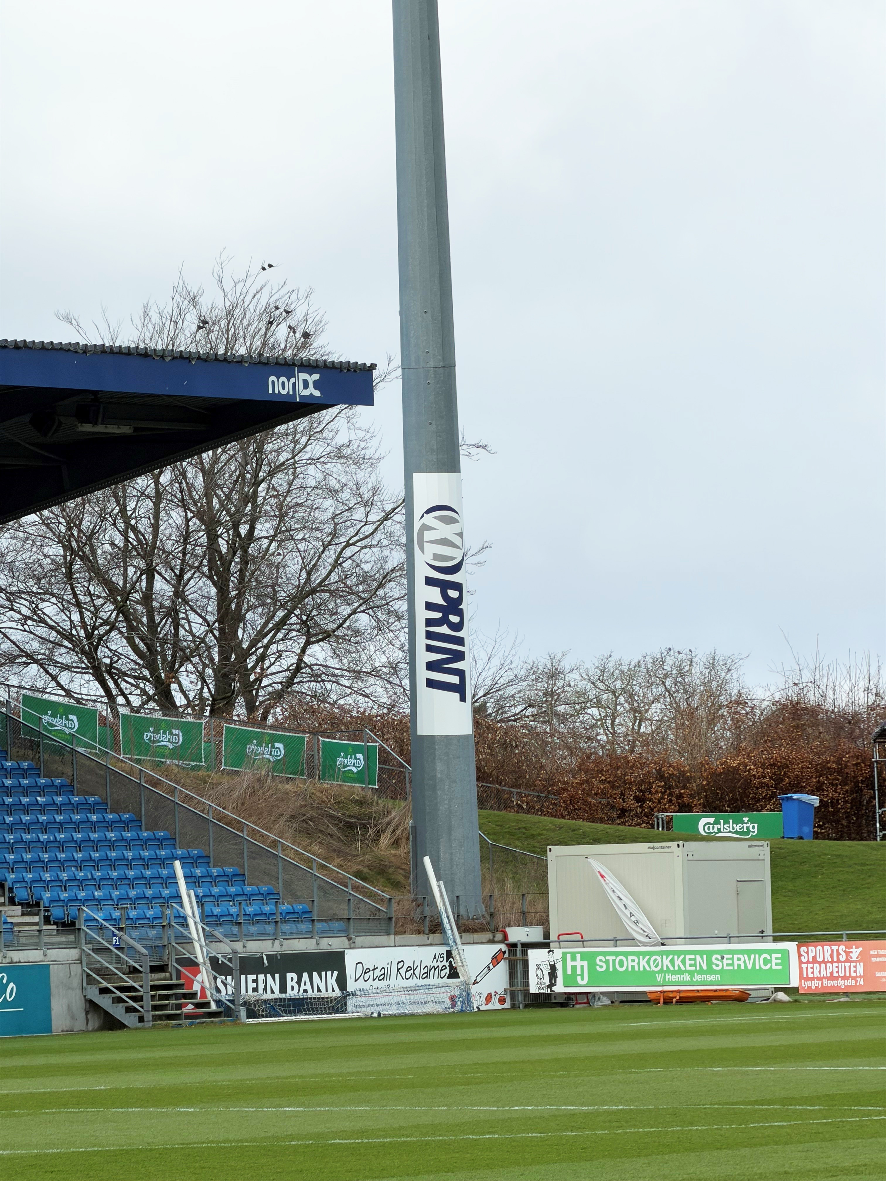 Fodboldstadion med grøn bane, blå tribunestole, og reklameskilte for blandt andet Carlsberg og Storkøkken Service under en grå himmel.