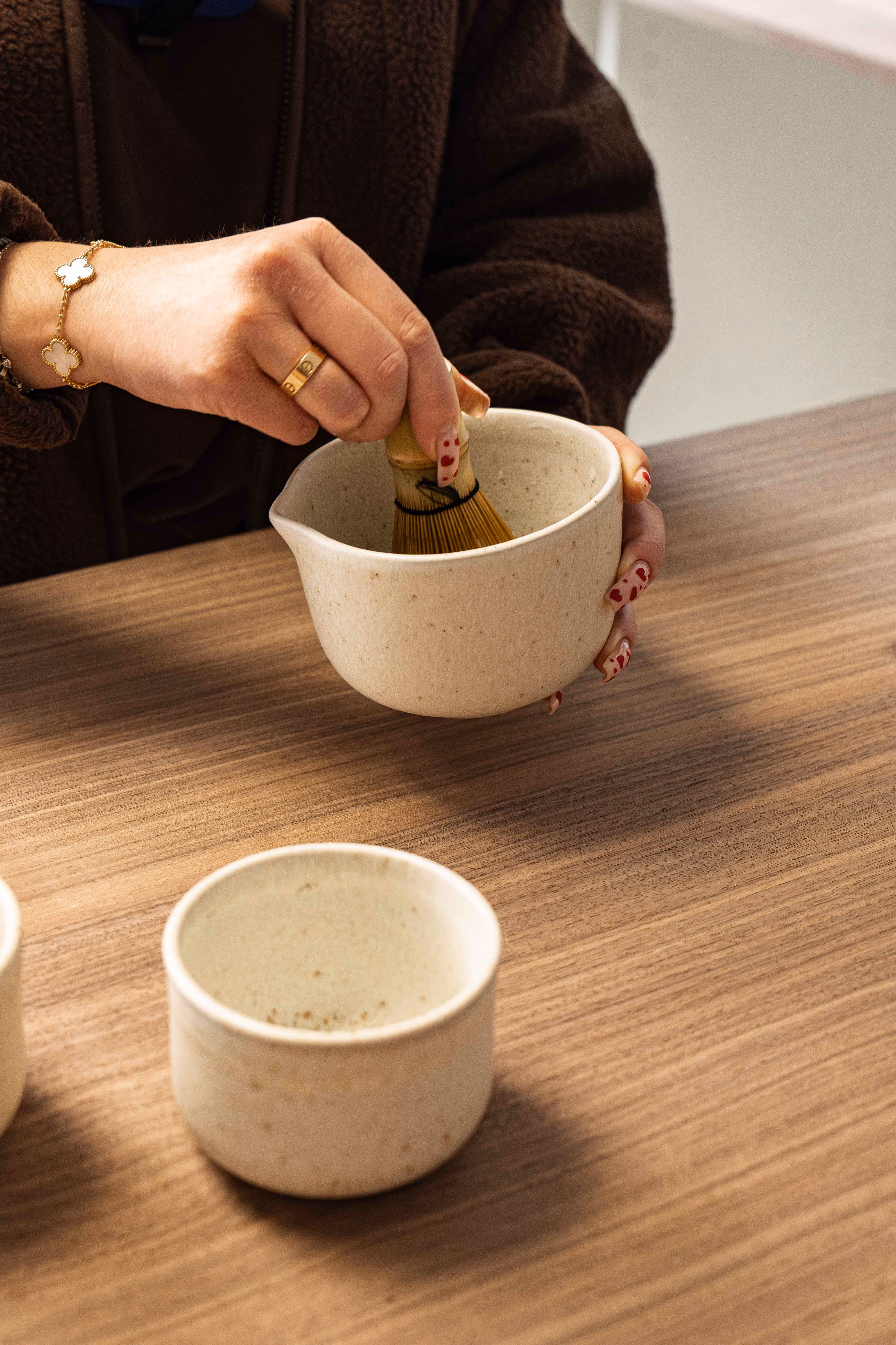 Main féminine tenant un fouet en bambou dans un bol beige sur une table en bois, prêt à préparer du thé.