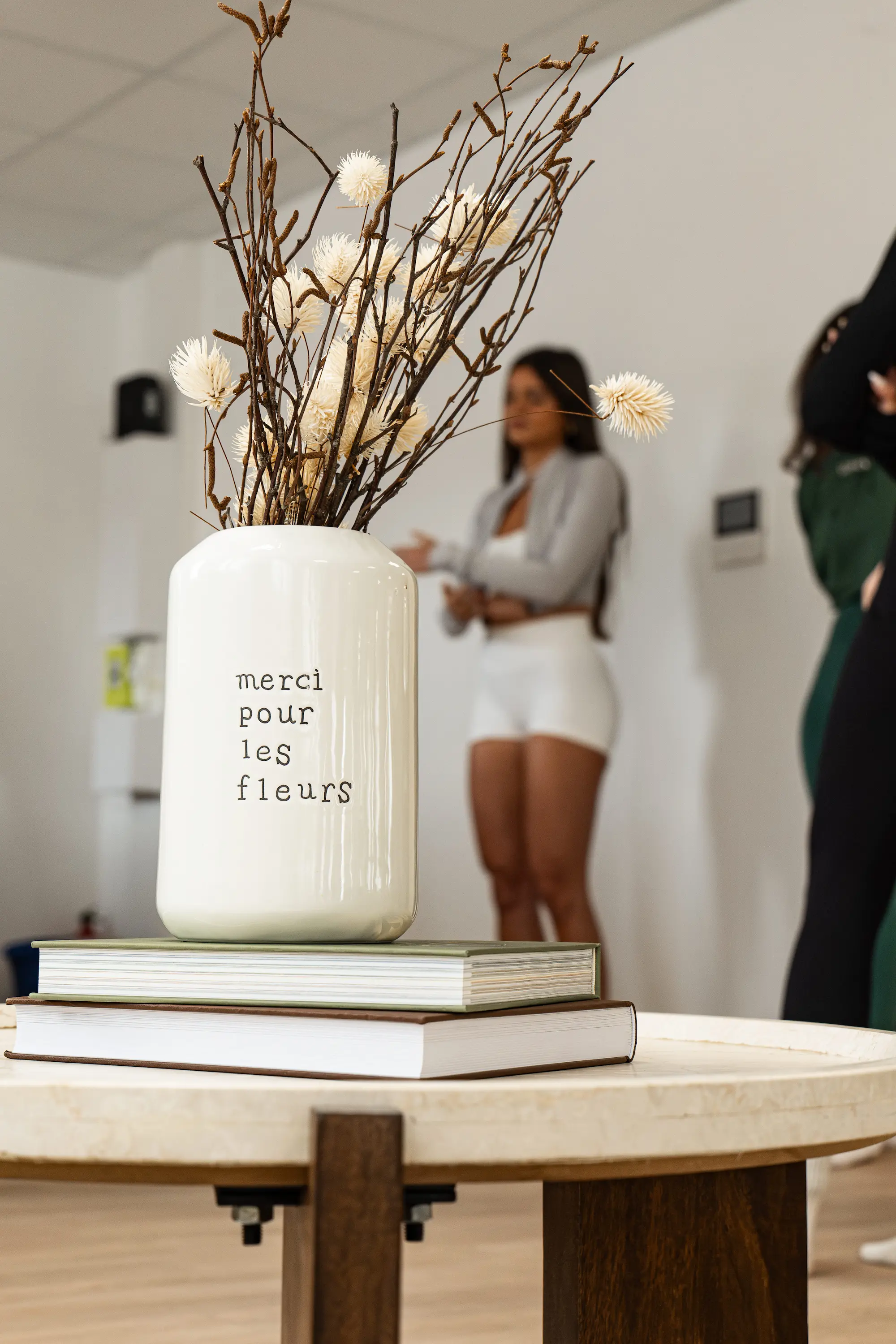 Vase blanc avec inscription « merci pour les fleurs » contenant des branches séchées et des fleurs posées sur deux livres sur une table, avec des personnes floues en arrière-plan.