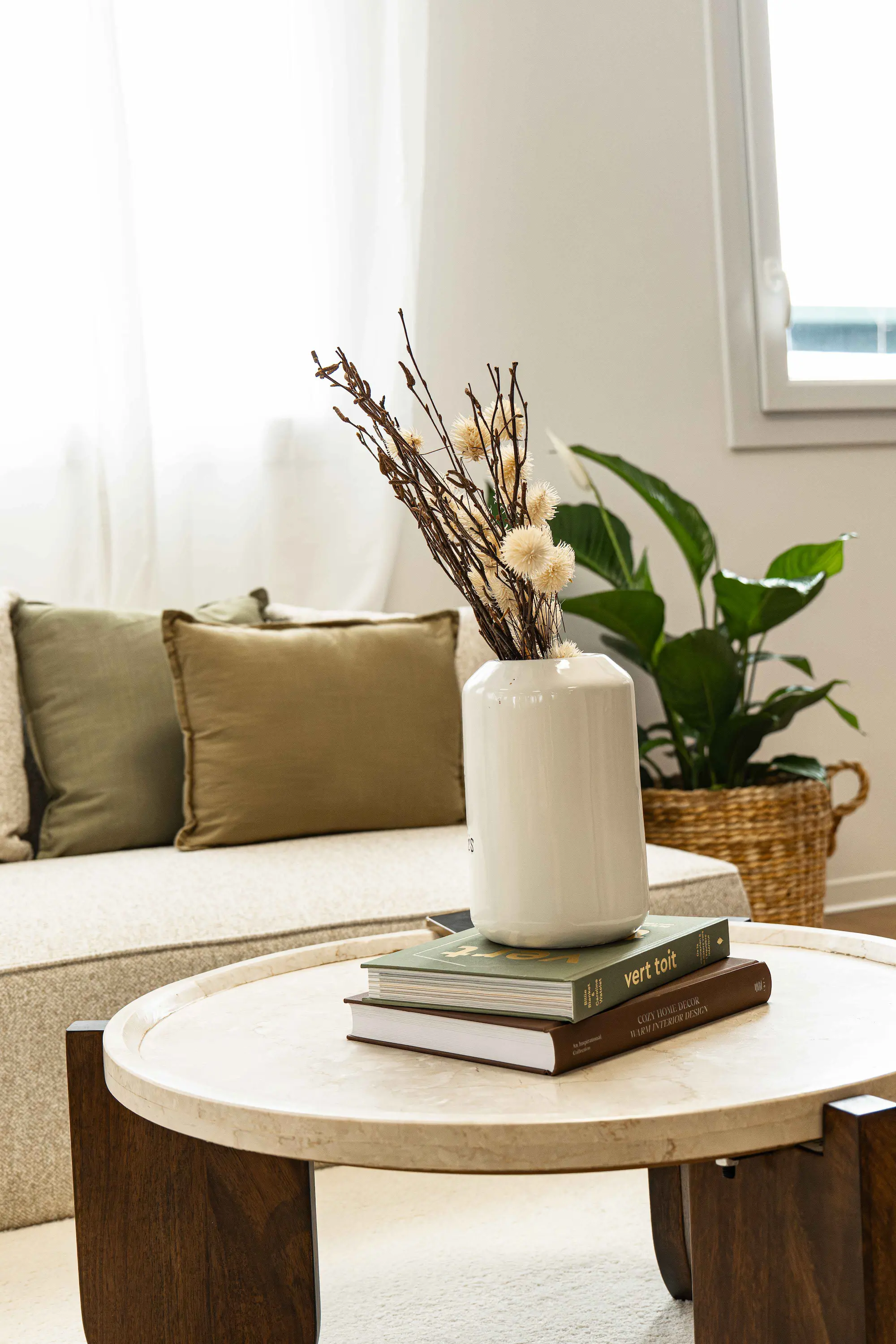 Table basse avec vase blanc contenant des branches et fleurs séchées, posée sur des livres, devant un canapé beige avec coussins verts et plante en panier tressé en arrière-plan.