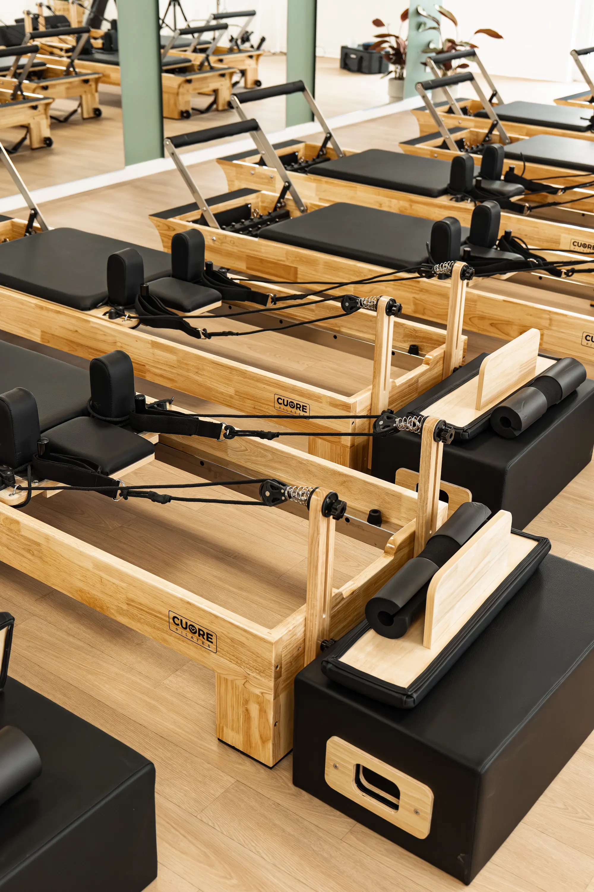 Salle de sport avec plusieurs reformers Pilates en bois clair et rembourrage noir alignés sur un sol en parquet clair près d'un miroir.