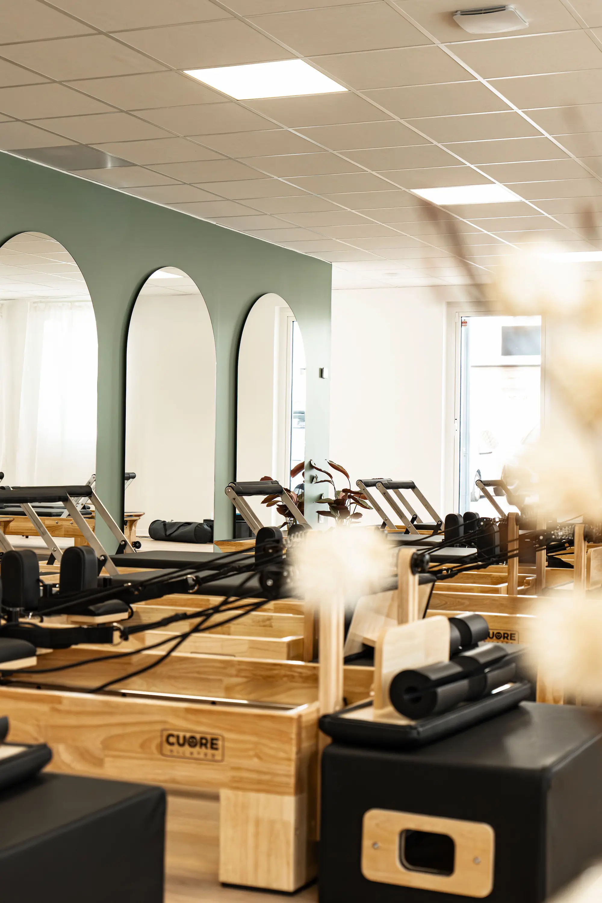 Salle de sport lumineuse avec des machines de Pilates en bois et miroirs arqués sur un mur vert.
