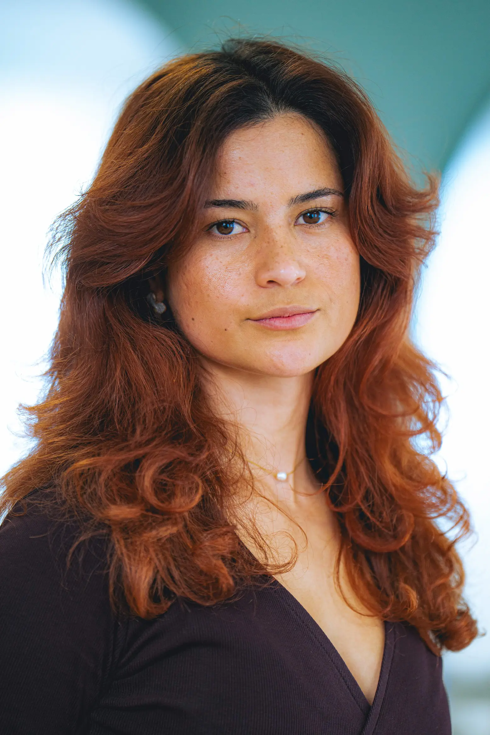 Portrait d'une femme avec de longs cheveux roux bouclés, portant un haut noir et un collier avec une perle.